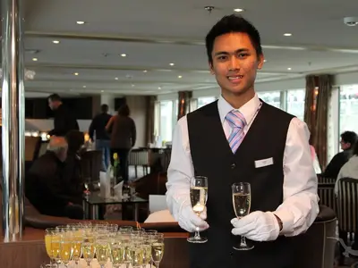 Ein lächelnder männlicher Crew-Mitarbeiter an Bord der MS Swiss Crystal in schwarzer Weste und weißen Handschuhen hält zwei Gläser Sekt. Im unscharfen Hintergrund sind weitere Gäste in der eleganten Lounge zu sehen.