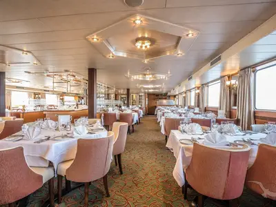 Ein geräumiges Restaurant an Bord der MS Swiss Crystal mit vielen weiß gedeckten Tischen und bequemen, braunen Stühlen. Die Fensterreihe bietet Tageslicht und Blick nach außen. Deckenleuchten und ein gemusterter Teppich ergänzen die elegante Atmosphäre.