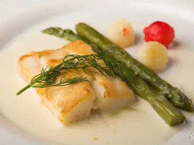 Ein Teller mit gebratenem Heilbutt, grünem Spargel, Kartoffelperlen und Dill in heller Soße, an Bord der MS Swiss Crystal.