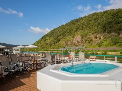 Blick auf das Sonnendeck der MS Swiss Crystal: Whirlpool, Liegestühle und Schirme. Im Hintergrund Fluss, bewaldeter Berg und blauer Himmel.