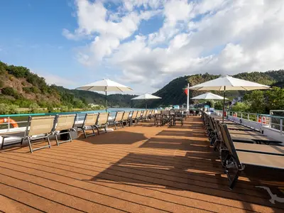 Einladendes Sonnendeck der MS Swiss Crystal mit Reihen von Liegestühlen und drei geöffneten Sonnenschirmen. Im Hintergrund eine grüne, hügelige Flusslandschaft unter blauem Himmel mit Wolken.