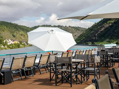 Das Sonnendeck der MS Swiss Crystal bietet bequeme Liegestühle und Tische unter Sonnenschirmen. Im Hintergrund sind ein Fluss und bewaldete Berge zu sehen.