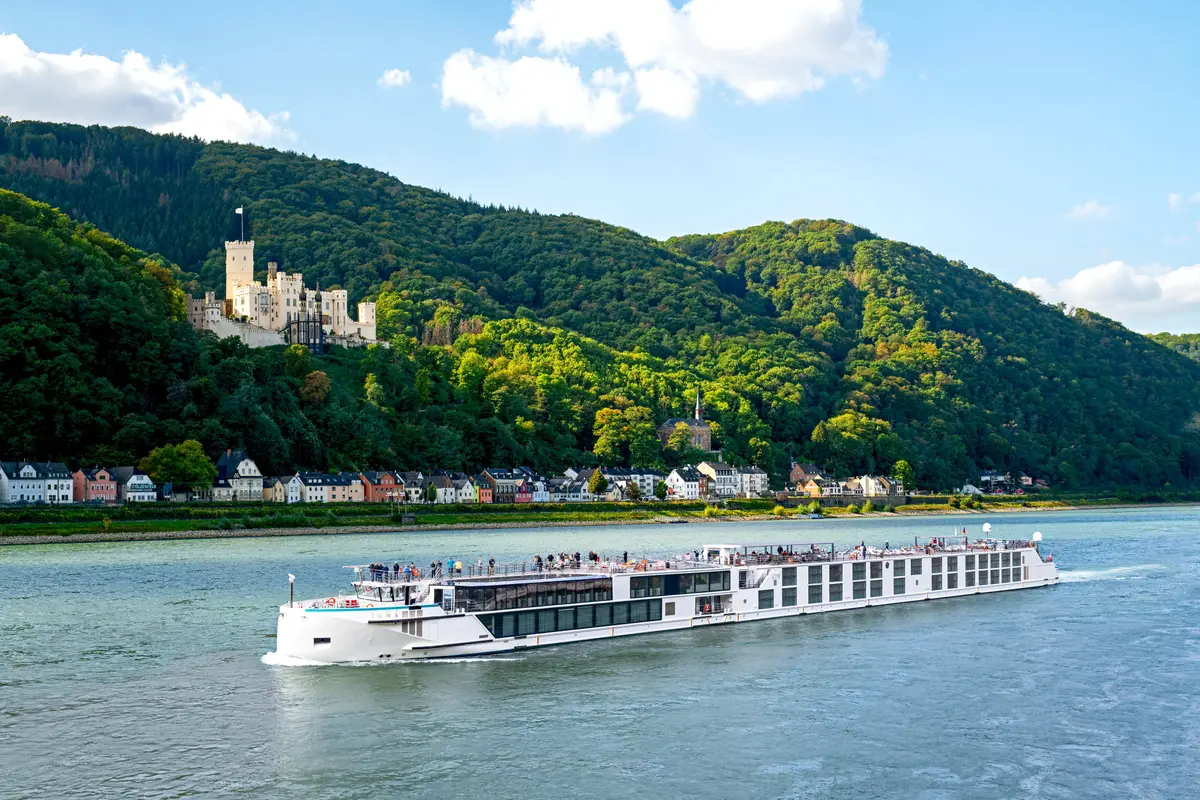 Die Riverside Debussy auf einem Fluss, umgeben von einem Schloss, einer Stadt und grünen Bergen unter blauem Himmel. Passagiere auf dem Sonnendeck.