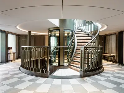 Modernes Atrium der Riverside Debussy mit eleganter Wendeltreppe, beleuchteten Stufen und einem runden Glasaufzug. Der Boden zeigt ein hell-dunkles Schachbrettmuster.