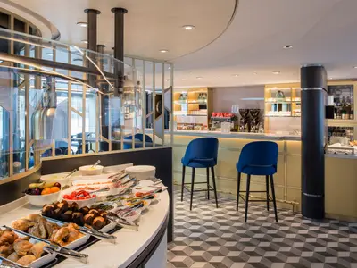 Modernes Buffet an Bord der Riverside Debussy. Gebäck, Obst, Salate und Aufschnitt sind appetitlich präsentiert. Im Hintergrund eine Bar mit blauen Hockern und Kaffeemaschinen.