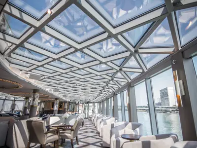 Stilvolle Lounge der Riverside Debussy mit Glasdach. Große Fenster bieten Flussblick auf Stadt und Brücke. Moderne, helle Sitzmöglichkeiten laden zum Verweilen ein.