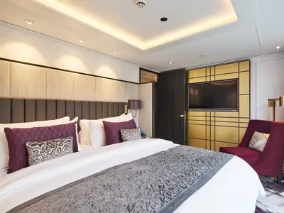 Ein elegant eingerichtetes Schlafzimmer in einer Suite an Bord der Riverside Debussy. Zu sehen sind ein großes Bett, eine gepolsterte Kopfzeile, eine goldene Wand mit Fernseher und ein roter Sessel.