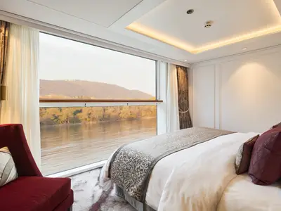 Suite an Bord der Riverside Debussy. Panoramafenster mit Blick auf einen Fluss und herbstliche Landschaft. Komfortables Doppelbett, roter Sessel, elegante Deckenbeleuchtung.