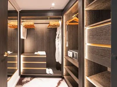 Ein luxuriöser, begehbarer Kleiderschrank in dunklem Holz mit goldenen Akzenten in einer Suite an Bord der Riverside Debussy. Helle Einbauleuchten erhellen den Bereich mit Kleiderbügeln, einem Bademantel, einem Safe und weissen Hausschuhen.