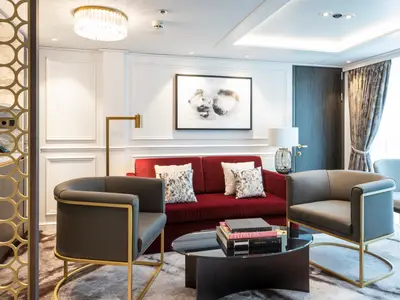 Luxuriöser Wohnbereich einer Suite an Bord der Riverside Debussy. Rotes Samtsofa, graue Sessel mit goldenem Gestell, Glastisch mit Büchern. Moderne Kunst an heller Wand, stilvolle Beleuchtung. Große Fenster.