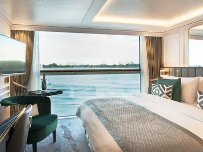 Luxuriöse Suite an Bord der Riverside Debussy mit grossem Bett, grünem Sessel und Schreibtisch. Die Panoramafenster bieten einen atemberaubenden Blick auf den Fluss.