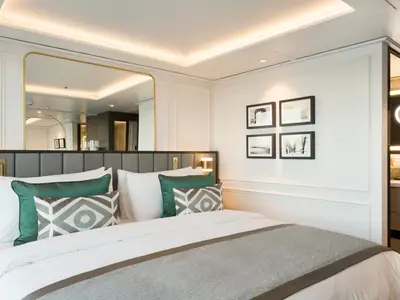 Luxuriöses Schlafzimmer in einer Suite der Riverside Debussy. Großes Bett mit grünen Kissen, goldgerahmter Spiegel und Wandbilder. Moderne, helle Einrichtung mit indirekter Beleuchtung.