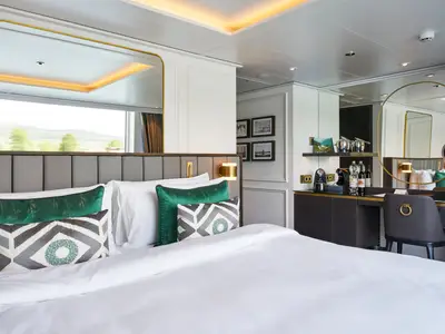 Geräumige Suite an Bord der Riverside Debussy. Großes Bett mit grünen Kissen. Rechts ein Schreibtisch mit goldgerahmtem Spiegel. Fensterblick auf grüne Landschaft.
