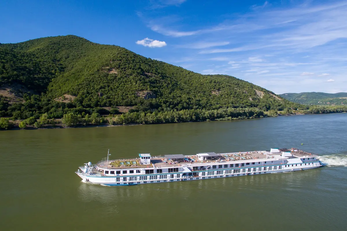 Blick aus der Vogelperspektive auf die Riverside Mozart auf einem Fluss. Das elegante Flussschiff gleitet vorbei an grünen Bergen unter blauem Himmel und hinterlässt eine weisse Heckwelle.