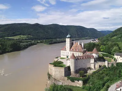 Panoramische Luftaufnahme der Burg Aggstein auf einem steilen Felsen ueber der Donau, mit bewaldeten Bergen und blauem Himmel. Eine malerische Kulisse entlang der Route der Riverside Mozart.