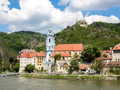 Blick auf Dürnstein an der Donau mit blauem Kirchturm, roten Dächern und Burgruinen auf grünem Berg. Gesehen vom Fluss aus an Bord der Riverside Mozart.