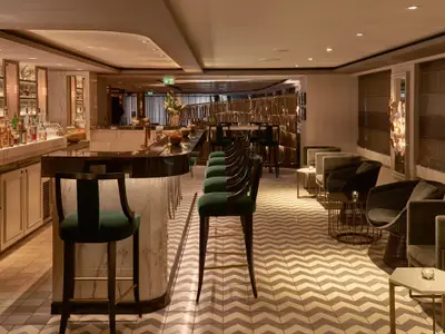 Die elegante Cove Bar an Bord der Riverside Mozart. Eine geschwungene Theke mit dunkelgrünen Samtbarhockern. Dahinter gemütliche Sesselbereiche auf einem Chevron-Teppich. Warme, luxuriöse Beleuchtung.