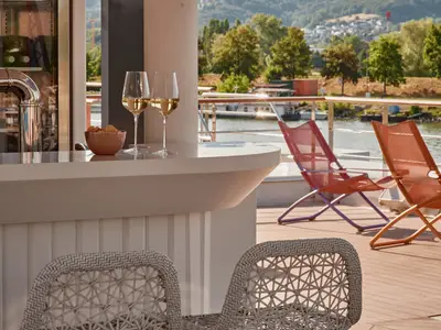 Zwei Gläser Weißwein und eine Schale Snacks stehen auf der weißen Bar an Bord der Riverside Mozart. Im Hintergrund entspannte Liegestühle und der Fluss.