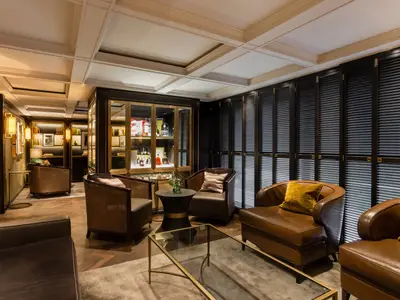 Elegante Lounge an Bord der Riverside Mozart. Komfortable Ledersessel, Glastische und eine beleuchtete Vitrine mit Flaschen sind zu sehen. Dunkles Holz, Bücherregale und Jalousien prägen das luxuriöse Interieur.