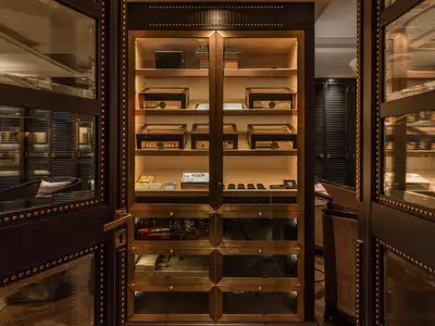 Ein luxuriöser, beleuchteter Zigarrenschrank mit Glasfächern und Messingrahmen, gefüllt mit Zigarren und Accessoires, an Bord der Riverside Mozart. Im Hintergrund sind Spiegelungen einer eleganten Lounge zu sehen.