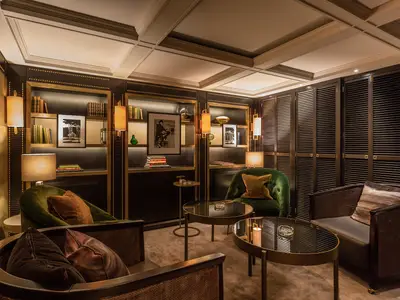 Eine elegante Lounge an Bord der Riverside Mozart mit dunklen Holzregalen, die mit Büchern und Dekorationen gefüllt sind. Mehrere grüne und braune Samtsessel stehen um runde Tische. Die Regale sind durch goldene Akzente und integrierte Beleuchtung hervorgehoben.