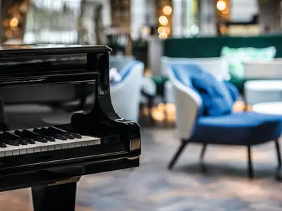 Schwarzes Klavier im Vordergrund der Lounge an Bord der Riverside Mozart. Unscharf im Hintergrund: blaue Sessel und grüne Couch in gemütlicher, hell beleuchteter Umgebung.