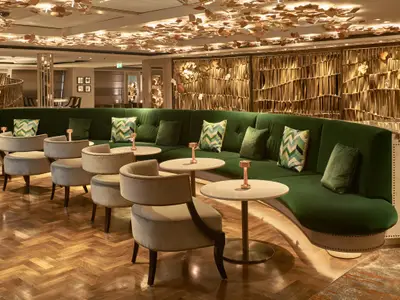 Luxuriöse Lounge an Bord der Riverside Mozart mit einer langen, grünen Sitzbank, beigen Sesseln und kleinen Tischen. Auffällige goldene Dekorationen an Decke und Wand. Holzboden.
