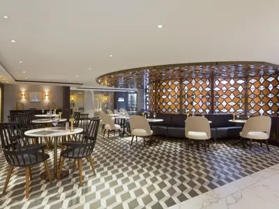 Bistro an Bord der Riverside Mozart mit modernen Tischen, schwarzen Stühlen und dunklen Sitzbänken. Der Boden hat ein geometrisches Muster. Eine rautenförmige orange-weiße Wandverkleidung ziert den Raum, beleuchtet von warmem Licht. Das Ambiente ist elegant und einladend, aber ohne Personen.