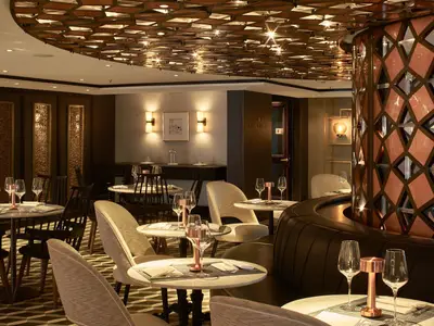 Innenansicht eines eleganten Restaurants an Bord der Riverside Mozart mit mehreren gedeckten Tischen, Weingläsern und modernen Leuchten. Eine dunkle Ledercouch ist rechts im Vordergrund sichtbar, umgeben von dunklen Holzvertäfelungen und einer dekorativen Decke.