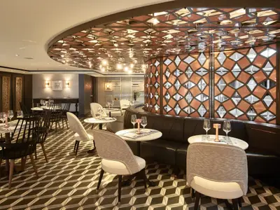 Stilvolles Bistro an Bord der Riverside Mozart. Moderne Tische, Stühle und Ledersofa. Decke und Wand mit beleuchteten, geometrischen Mustern. Gläser stehen bereit.