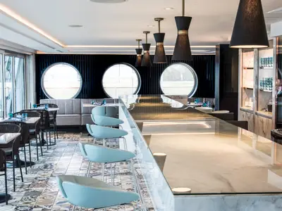 Modernes Restaurant "Blue Deli" an Bord der Riverside Mozart. Eine glänzende Bar mit hellblauen Hockern und Esstische sind zu sehen. Runde Fenster und elegante Einrichtung prägen den Raum.