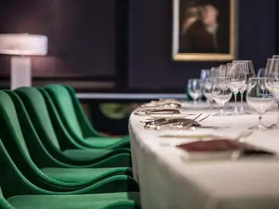 Elegantes Restaurant an Bord der Riverside Mozart. Festlich gedeckter Tisch mit Besteck, Gläsern und grünen Samtstühlen. Im Hintergrund eine Lampe und ein Gemälde.