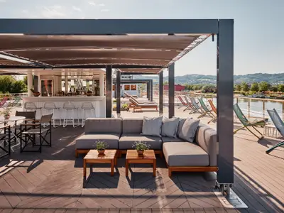 Helles Sonnendeck der Riverside Mozart mit Bar, Lounge-Sitzgelegenheiten und Liegestühlen. Blick auf den Fluss und bewaldete Hügel unter blauem Himmel.