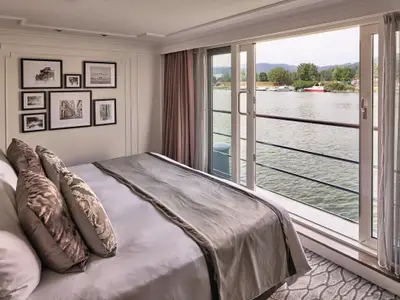 Eine elegant eingerichtete Suite an Bord der Riverside Mozart mit großem Doppelbett, vielen Kissen und einem Panoramafenster mit Blick auf einen Fluss und das Ufer. An der Wand hängen gerahmte Bilder.
