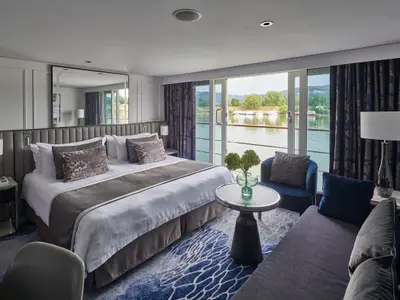 Luxuriöse Suite an Bord der Riverside Mozart. Doppelbett, Sitzecke und grosser Flussblick mit Booten und grünen Hügeln. Modernes Design.