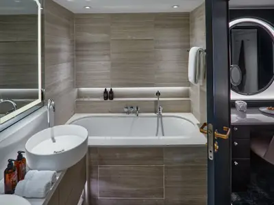 Ein modernes Badezimmer in einer Penthouse Suite an Bord der Riverside Mozart. Es zeigt einen ovalen Waschtisch, eine Badewanne, elegante Fliesen und eine beleuchtete Nische mit Pflegeprodukten.