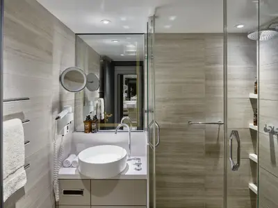 Helles, modernes Badezimmer an Bord der Riverside Mozart. Es zeigt einen Waschtisch mit ovalem Becken, einen beleuchteten Kosmetikspiegel und eine begehbare Dusche mit Regenduschkopf und Pflegeprodukten.