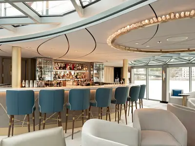 Ein moderner, luxuriöser Barbereich an Bord der Riverside Ravel. Eine geschwungene Bar mit blauen Barhockern und dahinter gut gefüllte Regale mit Spirituosen. Große Fenster bieten einen Blick auf eine Flusslandschaft mit Brücke und Gebäuden. Im Vordergrund sind bequeme cremefarbene Loungesessel zu sehen. Die Decke ziert eine beeindruckende ringförmige Lichtinstallation.