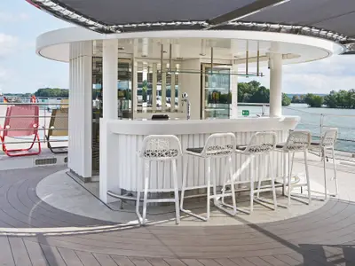 Eine helle, moderne Aussenbar auf dem oberen Deck der Riverside Ravel mit Barhockern. Im Hintergrund ist ein Fluss mit bewaldeten Ufern und Himmel zu sehen.