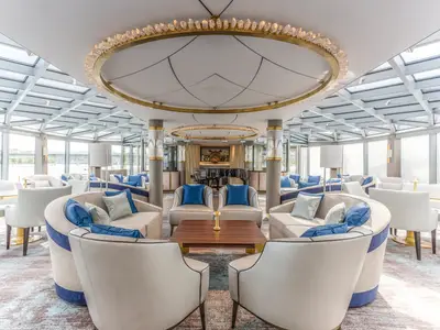 Die elegante Palm Court Lounge an Bord der Riverside Ravel. Luxuriöse Sitzgruppen in Blau und Beige unter einem Glasdach. Panoramafenster mit Flussblick und ein Flügel im Hintergrund.