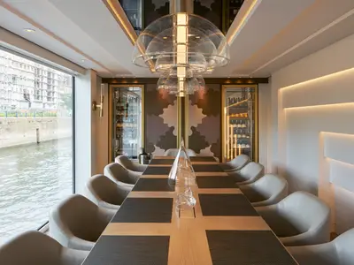 Der Vintage Room der Riverside Ravel: lange Tafel, helle Stühle, beleuchtete Weinregale. Großes Fenster mit Blick auf den Fluss und Uferbebauung.