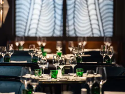 Eine Reihe elegant gedeckter Tische im Restaurant Waterside an Bord der Riverside Ravel, bereit für Gäste. Klare Weingläser und grüne Wassergläser schmücken die Tische, umgeben von dunklen Stühlen. Im Hintergrund sind Fenster mit dekorativen Vorhängen zu sehen, die eine angenehme Atmosphäre schaffen.