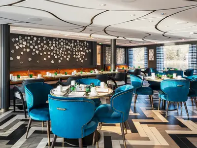 Innenansicht des modernen Restaurants Waterside an Bord der Riverside Ravel. Elegante blaue Samtstühle an runden Tischen mit Gedeck und Blick auf einen Fluss mit Vegetation.