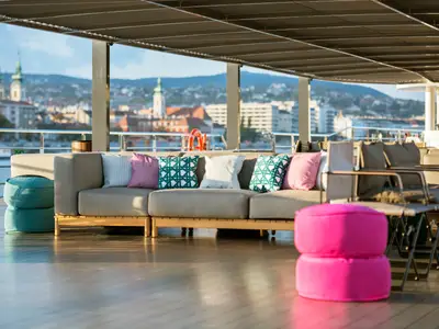 Einladendes Sonnendeck der Riverside Ravel mit komfortablen Outdoor-Sofas und bunten Kissen. Im Hintergrund eine malerische Stadtkulisse mit Kirchtürmen und Bergen.