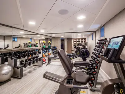 Ein modernes Fitnessstudio an Bord der Riverside Ravel. Es sind diverse Hantelstaender, ein Liegefahrrad und Rudergeraete zu sehen, reflektiert in grossen Spiegeln. Die Waende sind hell gehalten.