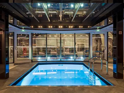 Blick auf den beleuchteten rechteckigen Innenpool an Bord der Riverside Ravel. Eine Metallleiter führt ins blaue Wasser. Die Decke und Wände sind aus Glas, was zu vielen Reflexionen führt.