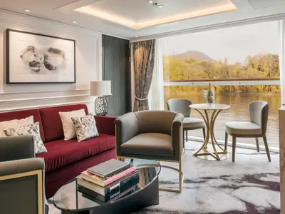 Einladende Suite an Bord der Riverside Ravel mit rotem Sofa, bequemen Sesseln und einem Fensterblick auf einen ruhigen Fluss und bewaldete Ufer. Ein Sektkühler steht bereit.