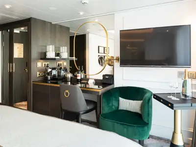 Blick in eine luxuriöse Suite der Riverside Ravel mit dunklem Holz, goldfarbenen Akzenten, Minibar, Schreibtisch und grünem Samtsessel. Ohne Personen.