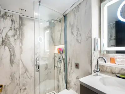 Ein elegantes Badezimmer an Bord der Riverside Ravel mit einer geräumigen Dusche und einem Waschtisch mit beleuchtetem Spiegel. Helle Marmorwände und moderne Chromarmaturen prägen das Design.