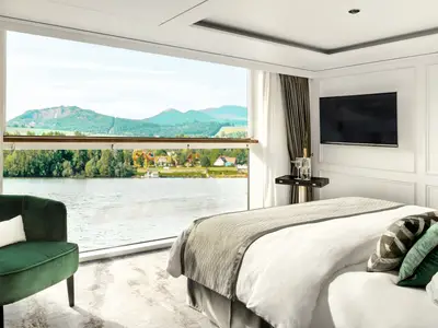Eine luxuriöse Suite an Bord der Riverside Ravel mit einem bequemen Bett, einem smaragdgrünen Sessel und einem grossen Fenster mit Blick auf einen Fluss und bewaldete Hügel.